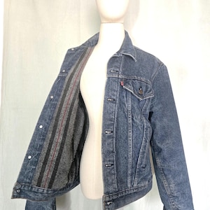 May include: A blue denim jacket with a button-up front and a collared neckline. The jacket has a gray and red striped lining and a small red tag on the chest pocket. The jacket is displayed on a mannequin.