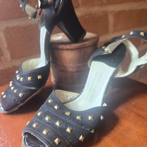 May include: A pair of black open-toe slingback heels featuring gold-tone pyramid studs across the front straps and ankle straps. The interior sole is labeled Saks Fifth Avenue. The shoes have a block heel and a light-colored inner lining.