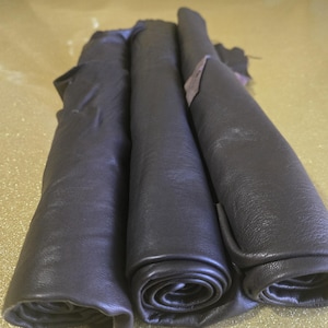 May include: Three rolls of dark gray leather, tightly curled, are displayed. The leather has a smooth, slightly shiny surface. The rolls are stacked next to each other, showcasing the texture and color variations of the material.