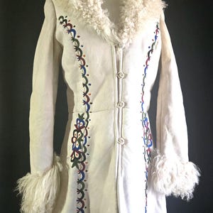 Pode incluir: Casaco creme com gola e punhos fofos em branco sujo. O casaco apresenta padrões florais bordados coloridos na frente e nas mangas. Possui três botões e um comprimento até a coxa.