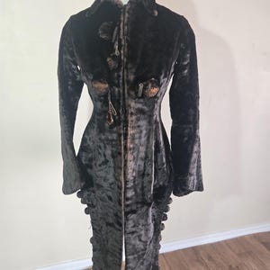 Puede incluir: Un abrigo vintage marrón oscuro con cuello alto y mangas largas. El abrigo presenta botones y adornos decorativos y se exhibe en un maniquí. El abrigo parece estar hecho de una tela de felpa texturizada.