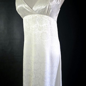 May include: A white, floor-length dress with a V-neckline and spaghetti straps. The dress features a floral pattern and is displayed on a mannequin. The straps are attached to the dress with metal rings.