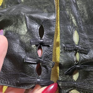 Op de afbeelding: Close-up van zwarte leren handschoenen met strikdetails. De handschoenen hebben uitgesneden accenten en kleine strikjes bij de openingen. Het leer lijkt getextureerd en de handschoenen staan tegen een gouden achtergrond.
