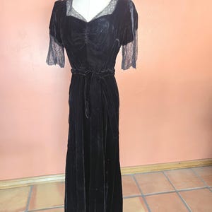 May include: A floor-length black velvet dress featuring short lace sleeves, a sweetheart neckline with lace trim, and a matching velvet tie belt at the waist. The bodice has a gathered center detail, and the skirt falls in a straight silhouette.