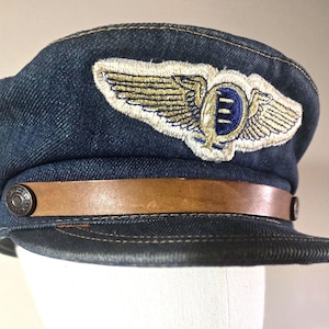 Può includere: Cappello da pilota in denim con fascia in pelle marrone e toppa decorativa con ali dorate e stemma blu. Il cappello ha una corona arrotondata e una visiera corta e rigida. Fissato con due bottoni metallici.