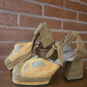 May include: A pair of olive green suede platform heels featuring a yellow snakeskin-textured toe box with light blue cutout accents. The shoes have a chunky block heel, a square toe, and a tie-up ankle strap closure with a light-colored interior lining.
