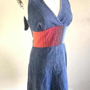 Puede incluir: Un vestido vaquero con cuello halter y escote en V con cintura entallada. La cintura presenta una tela a rayas vibrante en tonos rojos, rosas y negros. El vestido se exhibe en un maniquí.