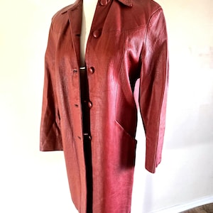 May include: A long, burgundy leather coat with a collared neckline and button-down closure. The coat features two front pockets and is displayed on a mannequin. The coat's color is a deep, reddish-brown.