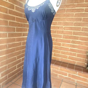 May include: A navy blue slip dress with delicate lace trim along the neckline, straps, and hem. The dress features a subtle bow detail at the bust and a flowing, knee-length silhouette. The dress is displayed on a mannequin.