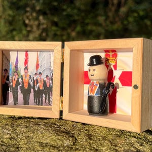 May include: A wooden hinged display box with two compartments. One side features a photograph of a parade, the other a wooden figure in a bowler hat and suit. The figure stands before a red and white background.