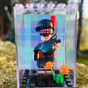 Könnte beinhalten: Eine Lego-Figur eines schottischen Dudelsackspielers in einem transparenten Display. Die Figur trägt einen schwarzen Hut, eine rote Jacke und einen Schottenkilt und hält eine Dudelsack. Orangefarbene Blumen befinden sich vor dem Gehäuse.