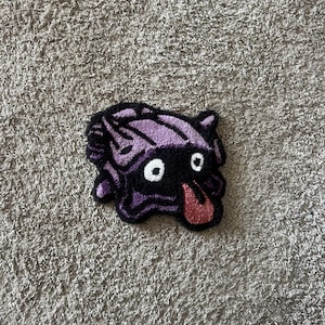 May include: A small, handmade rug in the shape of a cartoon monster. The rug features a black body with purple accents and a red tongue. The monster has white eyes and is laying down.