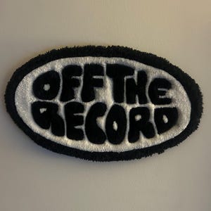 Può includere: Un cerotto ovale bianco e nero con il testo "OFF THE RECORD" in un font stilizzato.