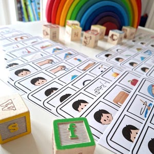Puede incluir: Un conjunto de juguetes educativos, que incluyen bloques de letras de madera y tarjetas ilustradas. Los bloques est&aacute;n pintados con letras y las tarjetas ilustradas presentan dibujos de objetos y personas. Un arco&iacute;ris de madera de colores se encuentra en el fondo.
