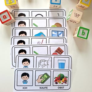Könnte beinhalten: Lernkarten mit Cartoon-Illustrationen und deutschen Wörtern. Die Karten zeigen Bilder eines Jungen, eines grünen Stifts, einer Kiwi, eines Fensters, einer Schokoladentafel, eines Wasserglases und Obst. Holzbuchstabenwürfel sind verstreut.