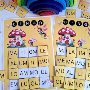 Puede incluir: Dos tarjetas de Bingo amarillas con ilustraciones de hongos y hadas. La palabra "BINGO" está en la parte superior. Pequeñas fichas amarillas con letras están esparcidas alrededor de las tarjetas. Un apilador de arcoíris de madera está en la parte superior.