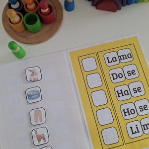 以下が含まれることがあります： 木製の虹、木製の人形、絵カード付きの本がセットになった学習教材。本にはウサギ、缶詰、ズボン、ラマ、飲み物の画像が掲載されています。ページには「Lama, Dose, Hase, Hose, Lim」と書かれています。