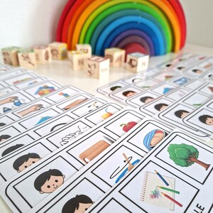 Puede incluir: Un conjunto de tarjetas did&aacute;cticas con ilustraciones y bloques de madera con s&iacute;mbolos. Un arco&iacute;ris de madera de colores est&aacute; en el fondo. Las tarjetas did&aacute;cticas presentan im&aacute;genes de objetos y actividades cotidianas.