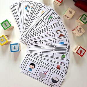 Puede incluir: Un conjunto de tarjetas did&aacute;cticas con contornos negros e ilustraciones, abanicadas sobre una superficie blanca. Las tarjetas presentan im&aacute;genes y palabras alemanas como "Ich", "Koche" y "Reis". Bloques de letras de madera con letras y n&uacute;meros coloridos est&aacute;n dispersos.
