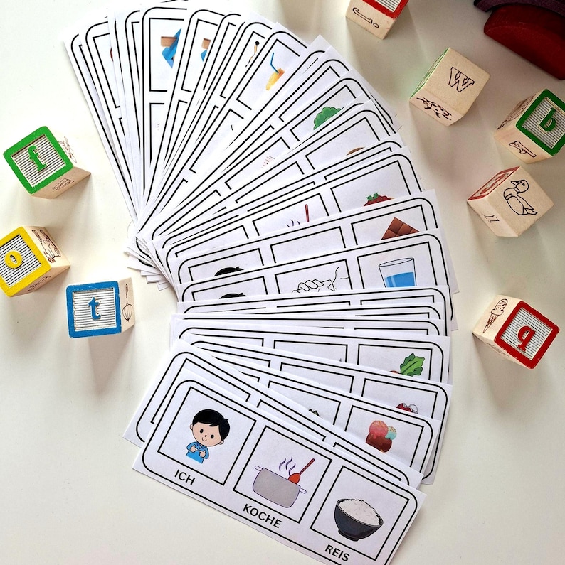 Puede incluir: Un conjunto de tarjetas did&aacute;cticas con contornos negros e ilustraciones, abanicadas sobre una superficie blanca. Las tarjetas presentan im&aacute;genes y palabras alemanas como "Ich", "Koche" y "Reis". Bloques de letras de madera con letras y n&uacute;meros coloridos est&aacute;n dispersos.