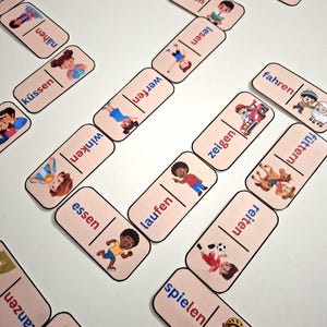 Puede incluir: Dominó con ilustraciones de niños realizando diversas acciones, como leer, correr y jugar. Cada ficha de dominó presenta una palabra en alemán y una imagen correspondiente. Los dominós están dispuestos sobre una superficie blanca.