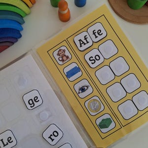 以下が含まれることがあります： 猿やソファなどの単語と画像が描かれたカードを使った子供向けの教育セット。追加の単語タイルと、さまざまな色の木製人形も含まれています。虹色の木製アーチが見えます。