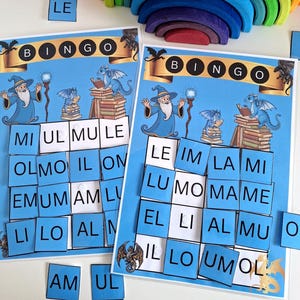 Op de afbeelding: Twee blauwe Bingo-spelborden met een thema van tovenaars en draken. Elk bord heeft een banner met de tekst "BINGO" en een raster van vierkanten met letters. Passende lettertegels liggen verspreid rond de borden. Een houten regenboog staat op de achtergrond.