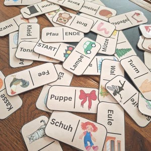 Puede incluir: Una colección de dominós de madera con ilustraciones y palabras en alemán. Los dominós presentan imágenes de un coche, un lazo, un gato y otros objetos. Las palabras incluyen "START", "ENDE" y otros términos alemanes.