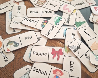 Könnte beinhalten: Eine Sammlung von Holzdomino-Steinen mit Illustrationen und deutschen Wörtern. Die Dominosteine zeigen Bilder von einem Auto, einer Schleife, einer Katze und anderen Objekten. Die Wörter umfassen "START", "ENDE" und andere deutsche Begriffe.
