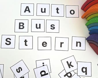 Könnte beinhalten: Weiße quadratische Buchstabenkacheln mit den Wörtern "Auto", "Bus" und "Stern". Zusätzliche lose Kacheln mit Buchstaben sind darunter verstreut. Ein farbenfroher Holz-Regenbogen-Stapler befindet sich in der oberen rechten Ecke. Lernspielzeug.
