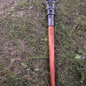 May include: A handheld torch featuring a long, cylindrical wooden handle attached to a dark metal cage head. The cage is constructed from vertical and horizontal metal bands, designed to hold a fuel source at the top of the wooden shaft.