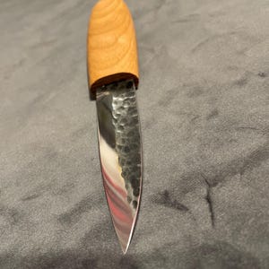 Puede incluir: Un pequeño cuchillo artesanal con un mango de madera clara y una hoja de metal martillado. La hoja es afilada y reflectante, con una punta. El mango es liso y redondeado. El cuchillo está diseñado para diversas tareas de corte. La hoja mide unos 10 cm.