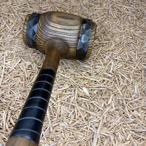 May include: A wooden mallet featuring a cylindrical head with dark brown wood grain and two black faceted bands on either side. The handle is made of brown wood with a black grip section featuring horizontal lines along the lower portion of the shaft.