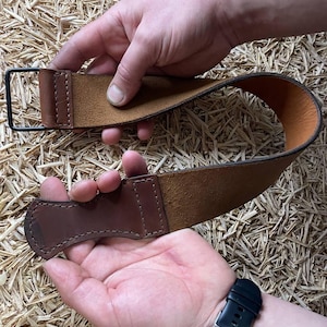 May include: A brown leather strop with a dark brown handle and a black metal loop. The strop is held by two hands against a background of straw.