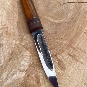 Pode incluir: Uma faca feita à mão com cabo de madeira e lâmina de aço polido. O cabo é de cor castanho escuro com uma faixa castanho escuro em torno do meio. A lâmina é afiada e tem uma ligeira curva.