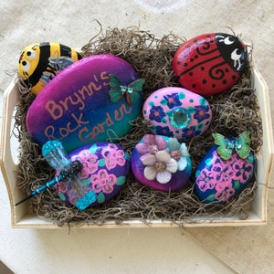 May include: A wooden tray filled with painted rocks. The rocks are decorated with various designs, including a ladybug, bee, flowers, and a dragonfly. One rock has the text "Brynn's Rock Garden". The rocks are nestled in dried moss.