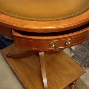 May include: A round wooden table with a leather top and a drawer. The table has a decorative edge and a brass handle on the drawer. The table is sitting on a wooden floor.