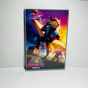 May include: A framed movie poster featuring a fox-like character in a blue and white outfit, holding a laser gun. The poster has a vibrant, colorful background with the text "THE SUPER MARIO GALAXY MOVIE" at the bottom.