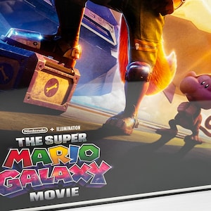 May include: Framed movie poster for "The Super Mario Galaxy Movie" featuring a character with a fox tail, a monkey-like creature, and a box with a wrench symbol. The poster has a black frame and a vibrant, colorful design.
