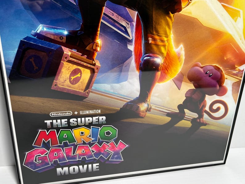 May include: Framed movie poster for "The Super Mario Galaxy Movie" featuring a character with a fox tail, a monkey-like creature, and a box with a wrench symbol. The poster has a black frame and a vibrant, colorful design.