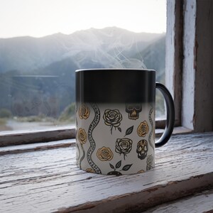 May include: A black heat-sensitive mug with a white design featuring a repeating pattern of yellow roses, snakes, and skulls. The mug is filled with a hot beverage, and steam is rising from the top.
