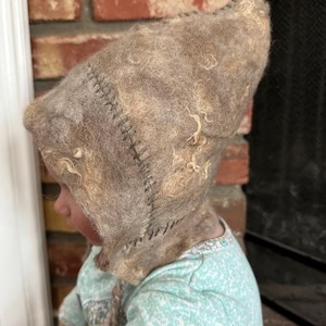 May include: A brown and white wool baby bonnet with a stitched seam. The bonnet is worn by a baby doll with a blue and white patterned dress.