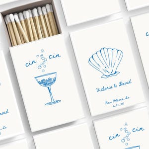 Könnte beinhalten: Ein Satz Streichholzschachteln mit einem blauen und weißen Design. Die Streichholzschachteln zeigen eine Muschel, ein Cocktailglas und den Text "cin cin". Der Text "Victoria & David" und "New Orleans, La 6.11.25" ist ebenfalls auf einer der Streichholzschachteln enthalten.