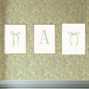 May include: Three framed art prints with light green designs against a floral wallpaper background. The prints feature a bow, the letter "A", and another bow. The frames are a light brown color.