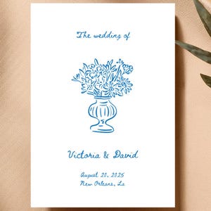 May include: A white wedding invitation with blue text. The text reads "The wedding of Victoria & David August 20, 2025 New Orleans, La". A blue line drawing of a vase with flowers is in the centre of the invitation.