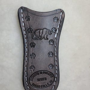 May include: A brown leather knife sheath with white stitching. The sheath has a bear and paw print design and the text "Richard M. Phillips Maker Idaho Falls, ID".