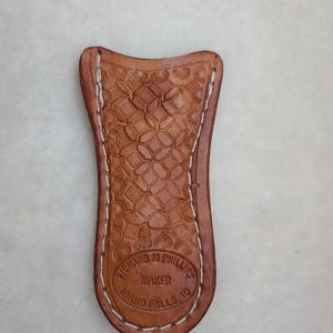 May include: A brown leather knife sheath with white stitching. The sheath has a decorative pattern and the text "Richard M. Phillips Maker Idaho Falls ID" is embossed on the bottom.
