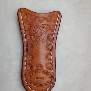 May include: A brown leather knife sheath with a stitched edge and an embossed trout design. The sheath is stamped with "Richard M. Phillips Maker Idaho Falls, ID."