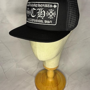 May include: Black trucker hat with a flat brim and mesh back. The front features a white embroidered patch with the text "CHROME HEARTS HOLLYWOOD, USA" and a decorative logo.