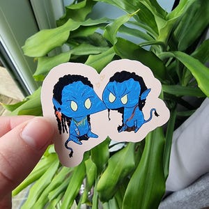 May include: A sticker featuring two blue cartoon characters with black hair and yellow eyes. The characters are touching noses and have small tails. The sticker is on a light pink background with two small hearts. The sticker is held in front of a green plant.
